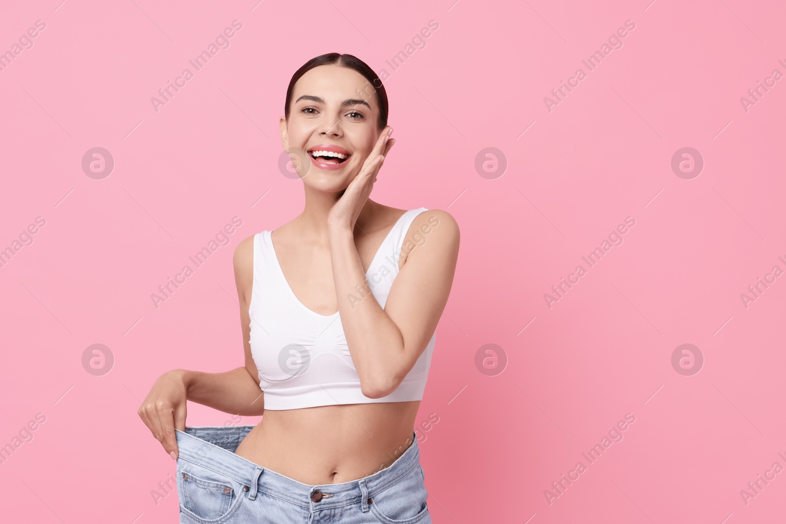 Photo of Diet and weight loss concept. Happy young woman in big jeans showing her slim body on pink background. Space for text