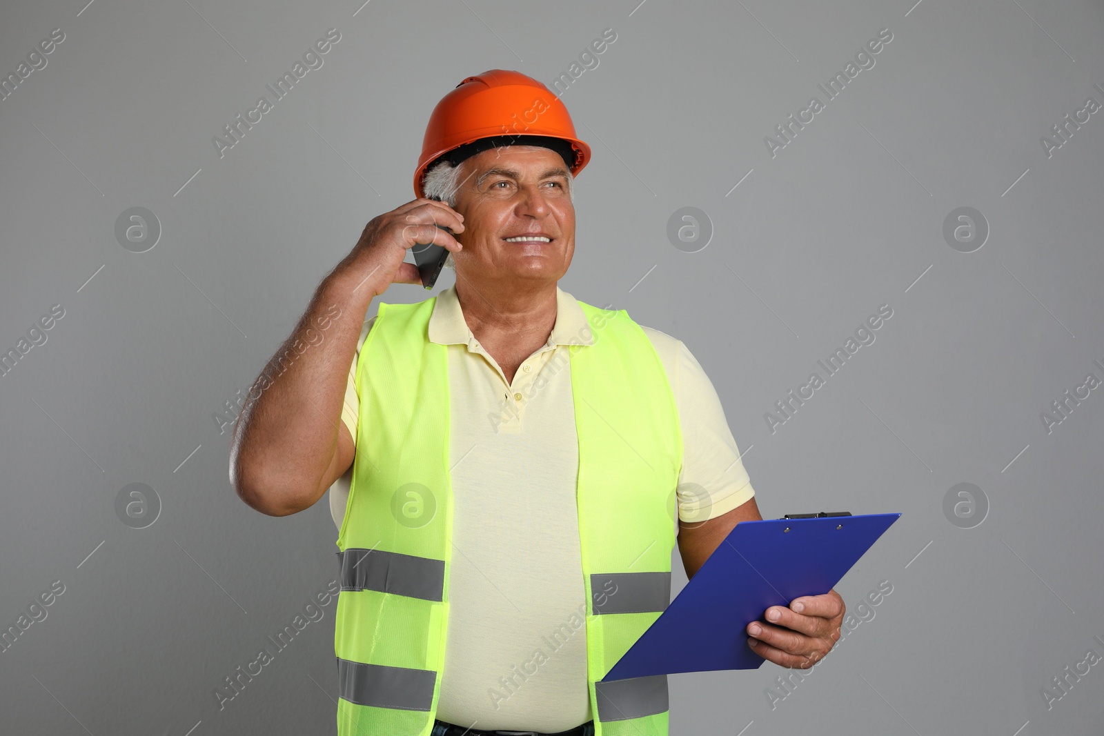 Photo of Engineer in hard hat with clipboard talking on smartphone against grey background