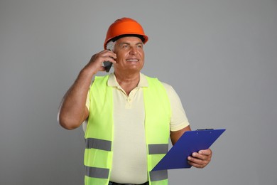 Photo of Engineer in hard hat with clipboard talking on smartphone against grey background