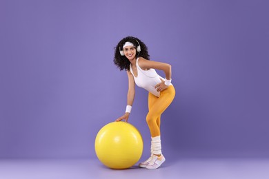 Photo of Aerobics. Happy woman in headphones with fitness ball on purple background