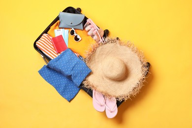 Open suitcase with traveler's belongings on yellow background, top view