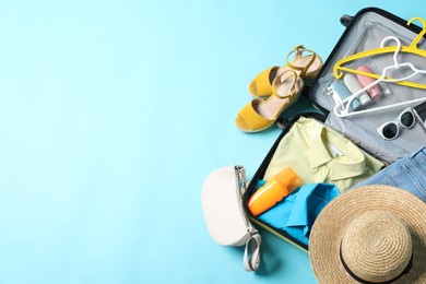 Open suitcase with traveler's belongings on light blue background, flat lay. Space for text