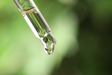 Photo of Essential oil dripping from pipette against blurred green background, closeup. Space for text