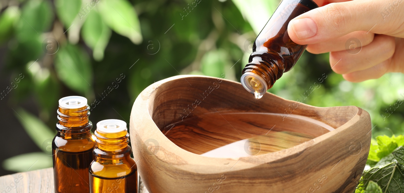 Photo of Woman dripping essential oil into bowl of water against blurred green background, closeup. Banner design