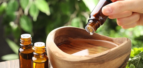 Photo of Woman dripping essential oil into bowl of water against blurred green background, closeup. Banner design