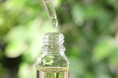 Photo of Essential oil dripping from pipette into bottle against blurred green background, closeup. Space for text