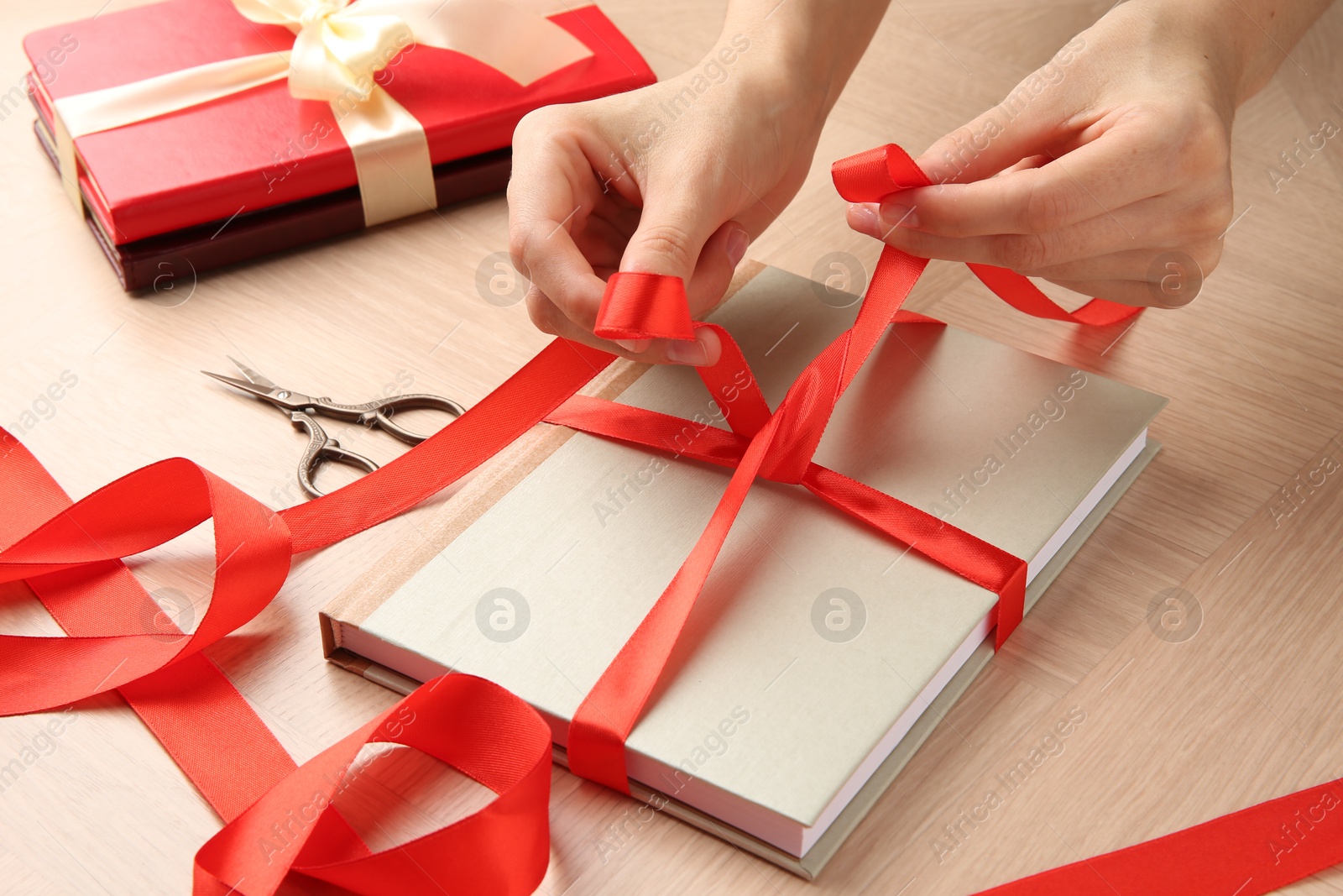 Photo of Woman tying ribbon on book at wooden table, closeup. Preparing gift