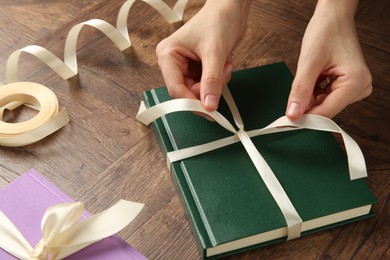 Photo of Woman tying ribbon on book at wooden table, closeup. Preparing gift