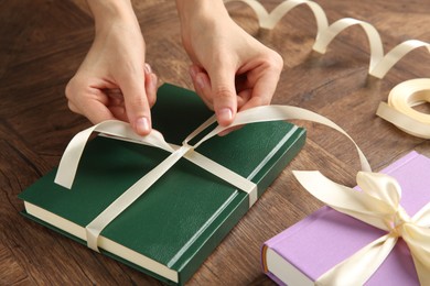 Woman tying ribbon on book at wooden table, closeup. Preparing gift