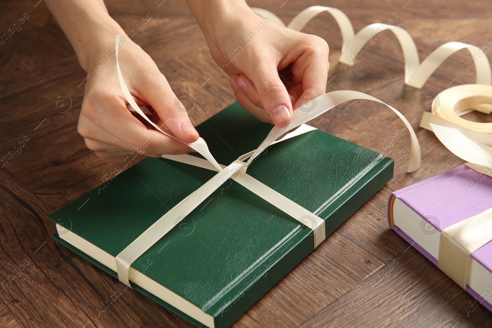 Photo of Woman tying ribbon on book at wooden table, closeup. Preparing gift