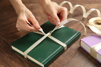 Photo of Woman tying ribbon on book at wooden table, closeup. Preparing gift