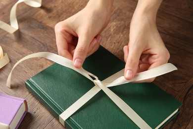 Photo of Woman tying ribbon on book at wooden table, closeup. Preparing gift