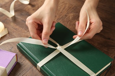 Photo of Woman tying ribbon on book at wooden table, closeup. Preparing gift