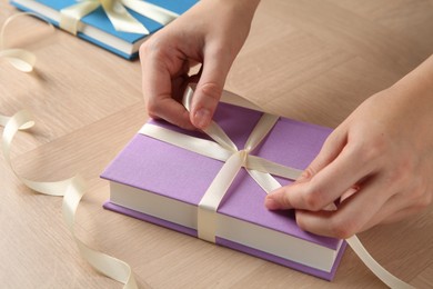 Woman tying ribbon on book at wooden table, closeup. Preparing gift