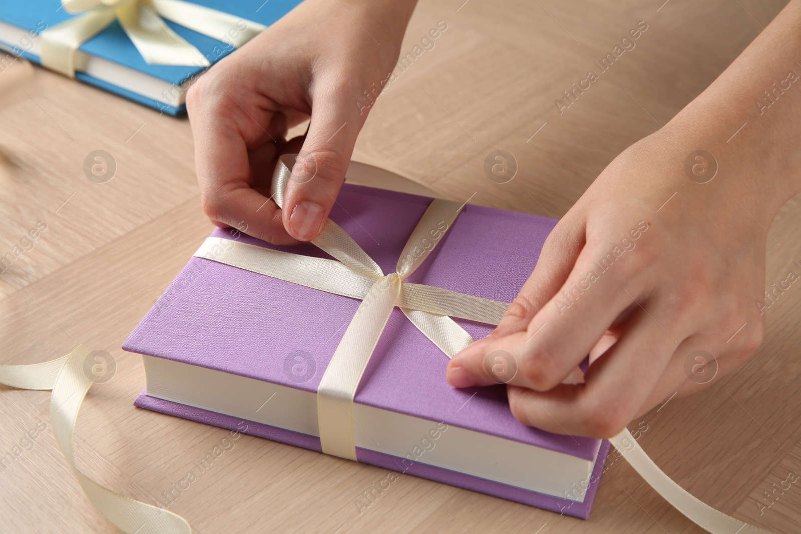 Photo of Woman tying ribbon on book at wooden table, closeup. Preparing gift