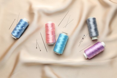 Photo of Spools of sewing threads, pins and needles on beige cloth, top view