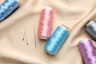 Photo of Spools of sewing threads, pins and needles on beige cloth, top view