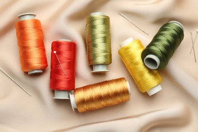 Photo of Spools of sewing threads, pins and needles on beige cloth, top view