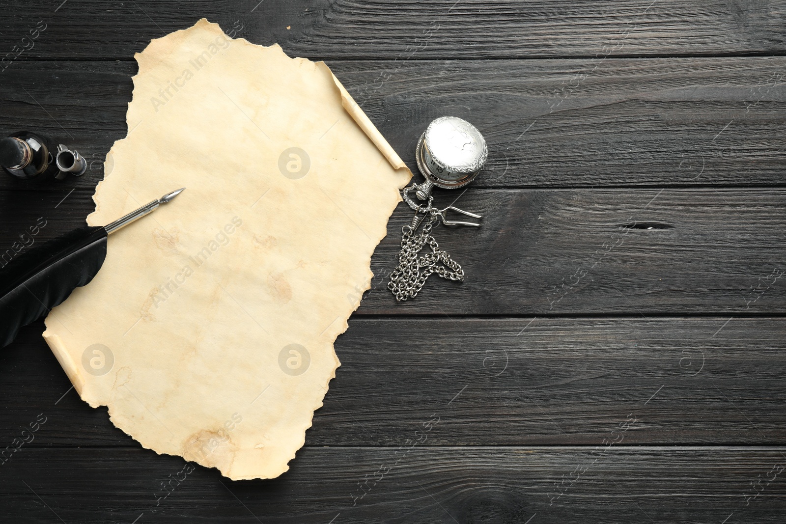 Photo of Sheet of old parchment paper, feather, inkwell and pocket chain clock on black wooden table, top view. Space for text