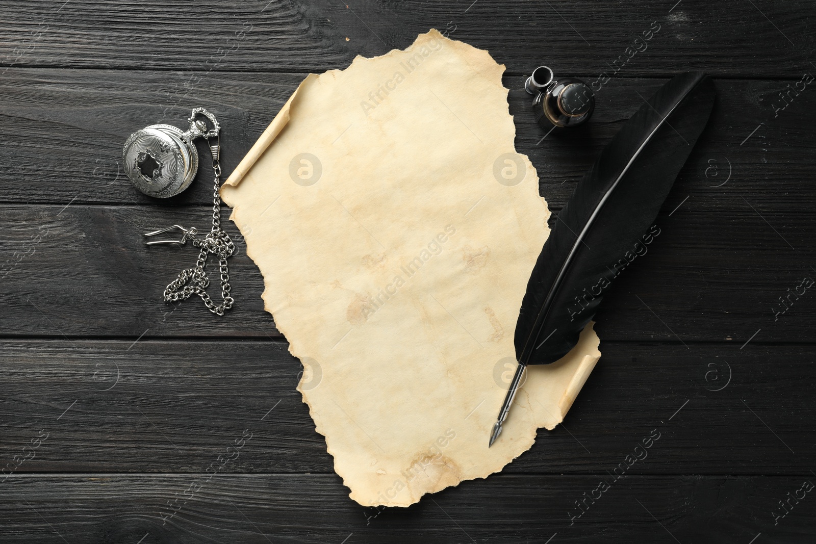 Photo of Sheet of old parchment paper, feather, inkwell and pocket chain clock on black wooden table, top view