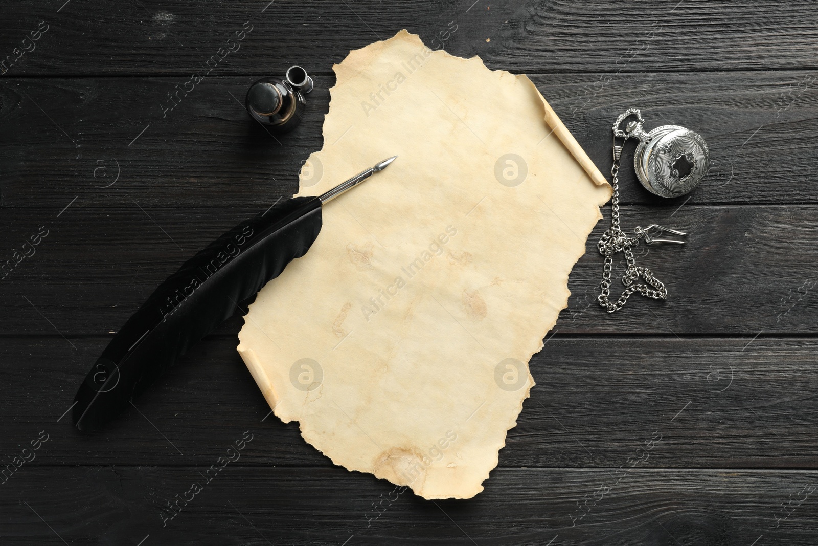 Photo of Sheet of old parchment paper, feather, inkwell and pocket chain clock on black wooden table, top view