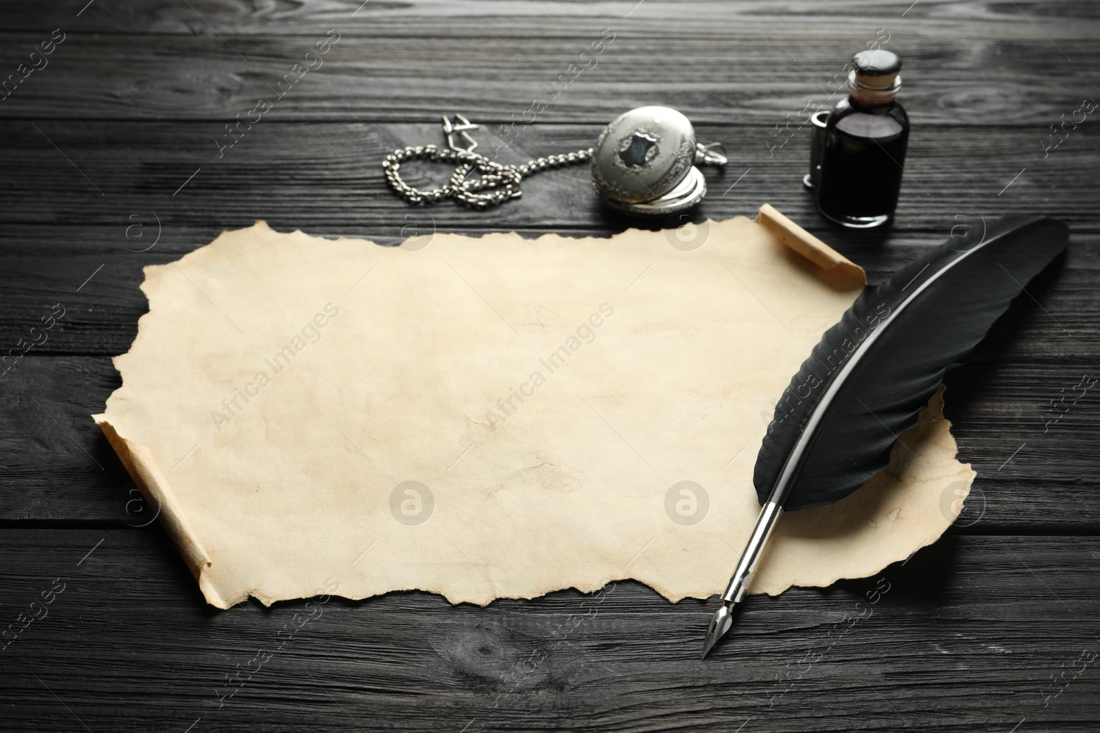 Photo of Sheet of old parchment paper, feather, inkwell and pocket chain clock on black wooden table