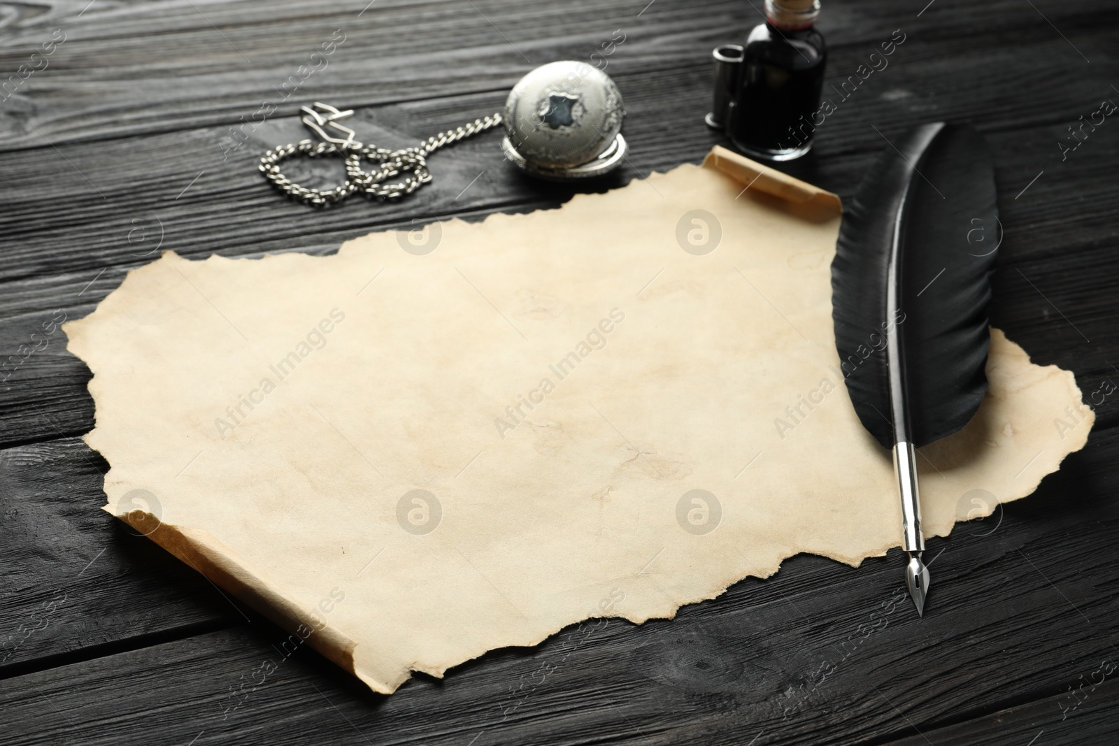 Photo of Sheet of old parchment paper, feather, inkwell and pocket chain clock on black wooden table