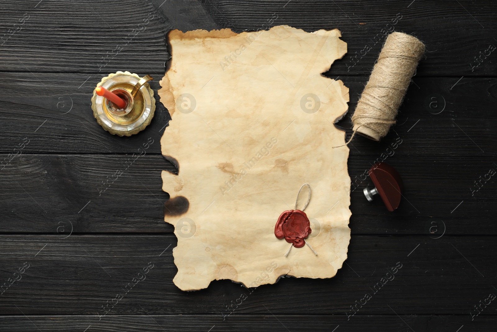 Photo of Sheet of old parchment paper with wax stamp, twine and candle on black wooden table, top view