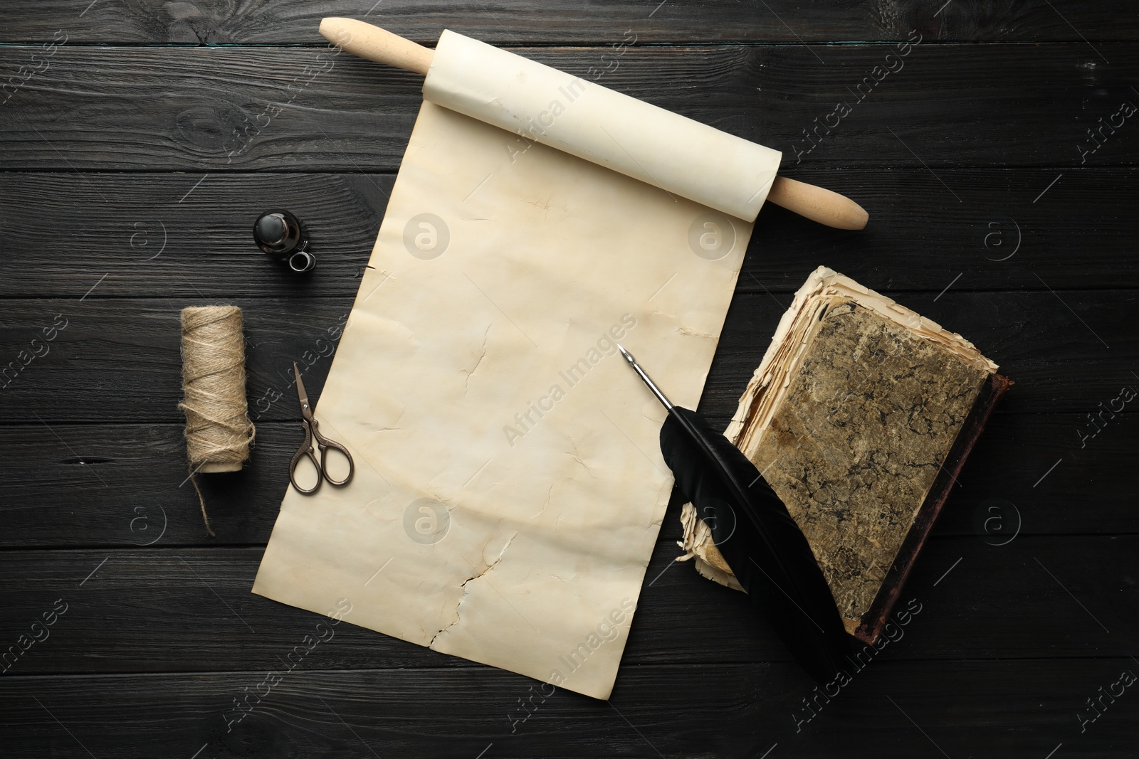 Photo of Sheet of old parchment paper, inkwell, scissors, vintage book, feather and twine on black wooden table, top view