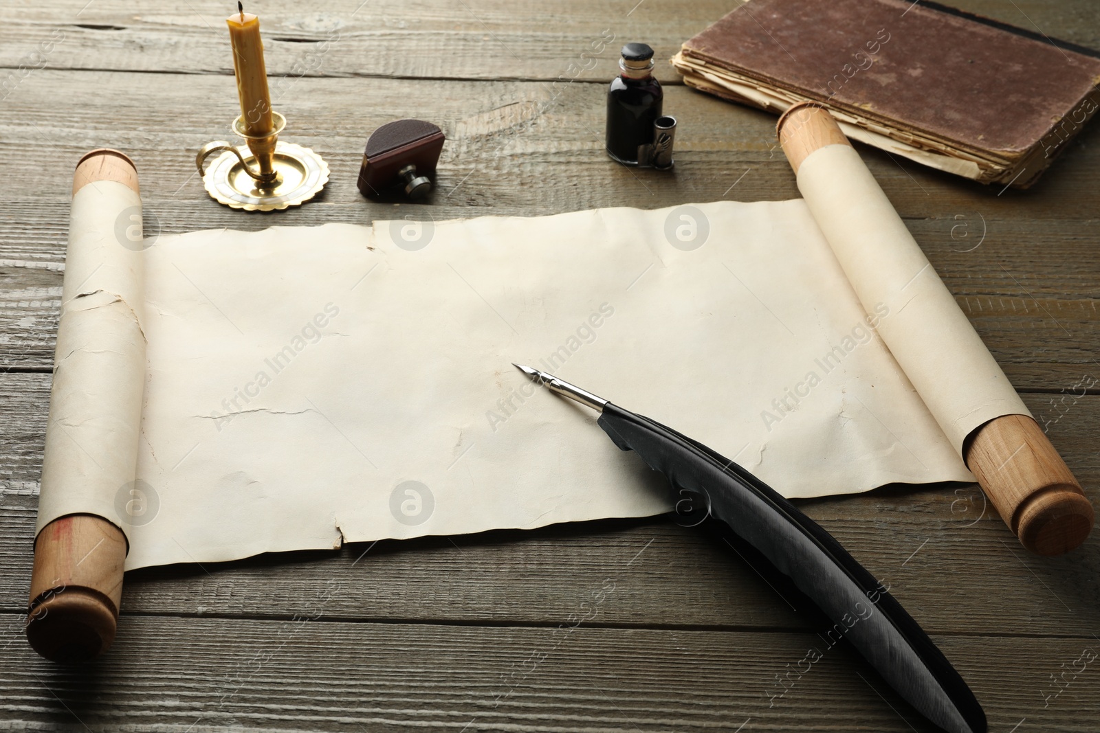 Photo of Sheet of old parchment paper, wax stamp, black feather, inkwell, candle and vintage book on wooden table