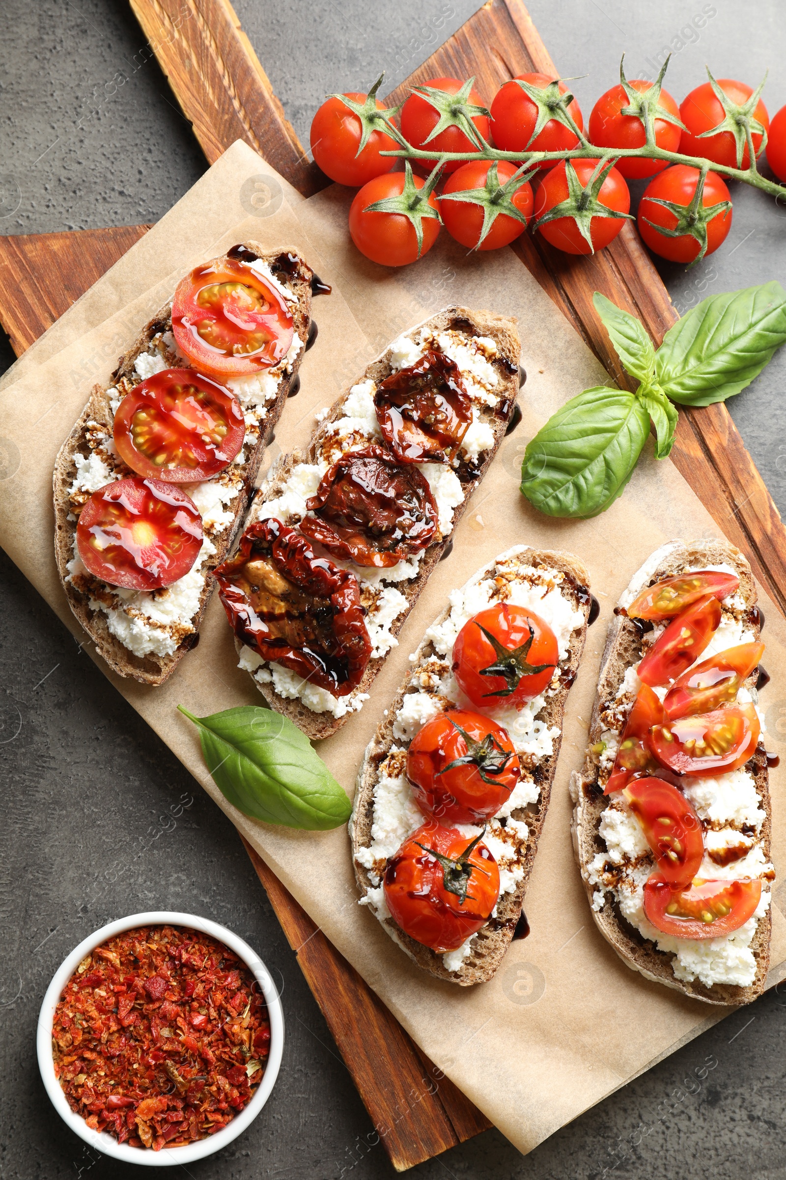 Photo of Delicious ricotta bruschettas with sun dried tomatoes, milled chili pepper and basil on grey textured table, flat lay