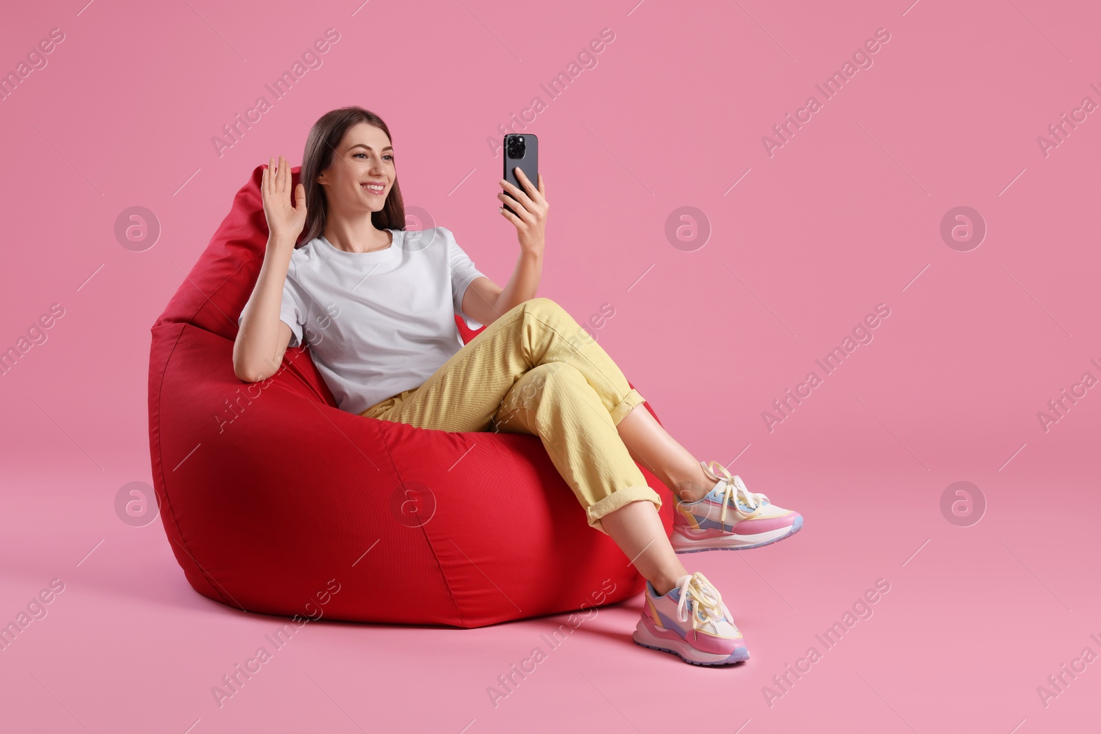 Photo of Smiling woman with smartphone having online meeting on red bean bag chair against pink background