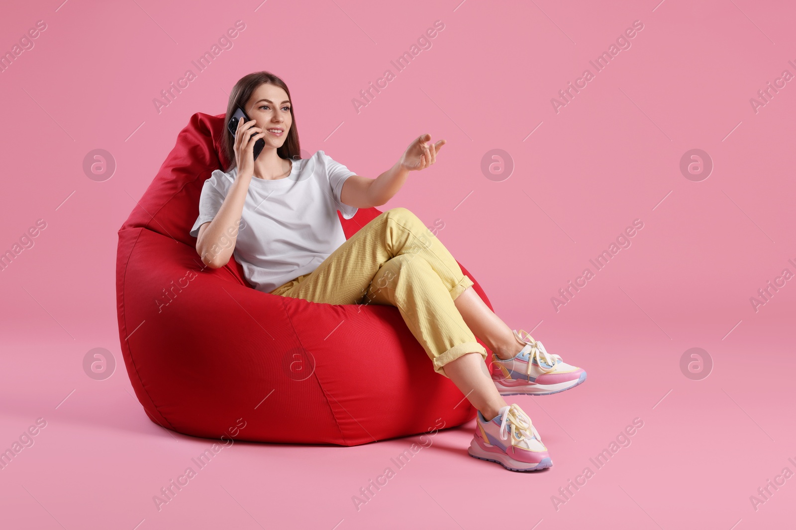 Photo of Smiling woman talking on smartphone while sitting on red bean bag chair against pink background