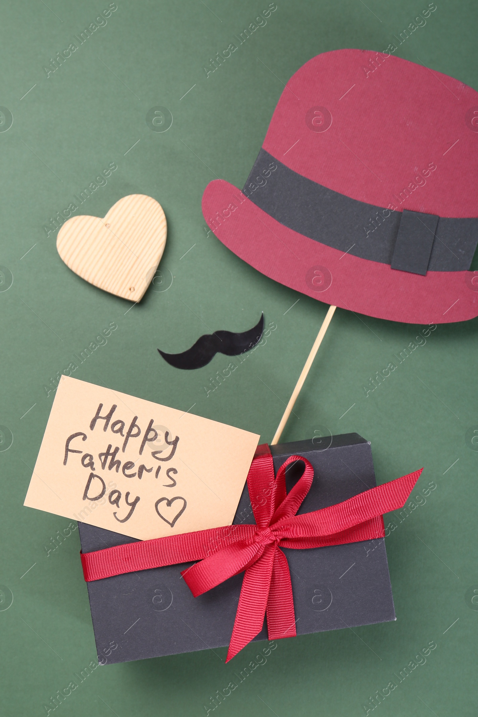 Photo of Card with phrase Happy Father's Day, gift box, heart, moustache and paper hat on green background, flat lay