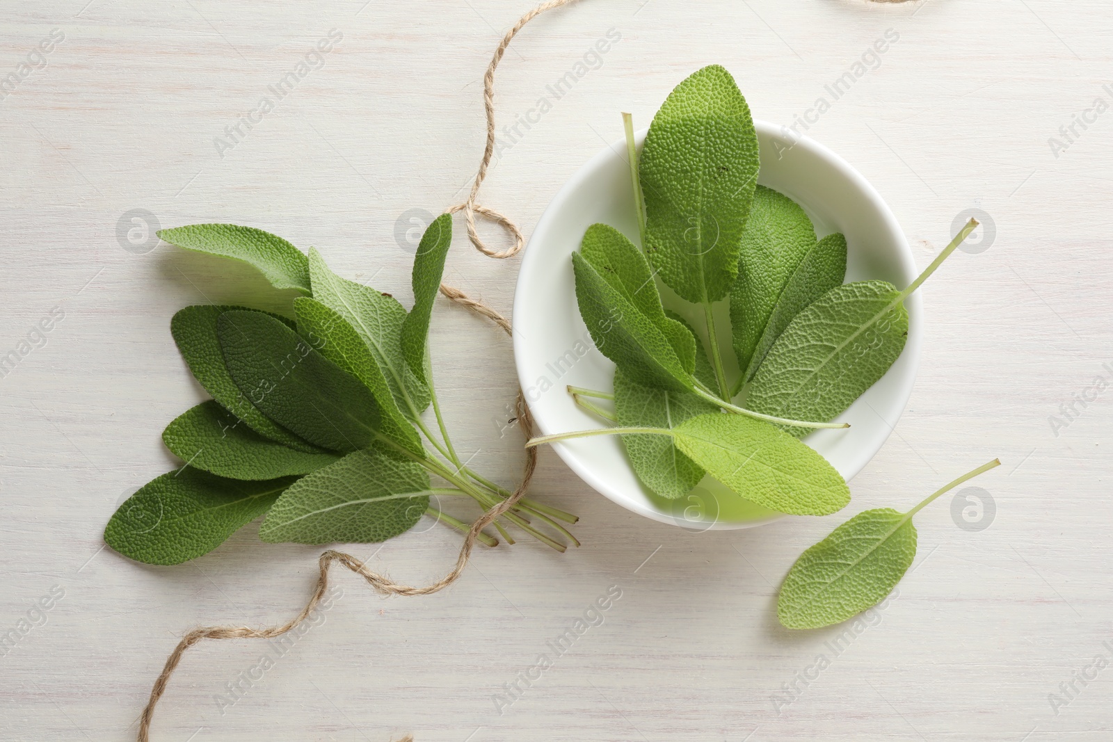 Photo of Fresh sage leaves in bowl and thread on light wooden table, top view