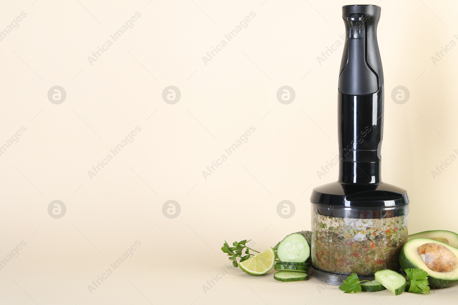 Photo of Hand blender with mixture of ingredients and fresh products on beige background, space for text