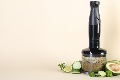 Hand blender with mixture of ingredients and fresh products on beige background, space for text