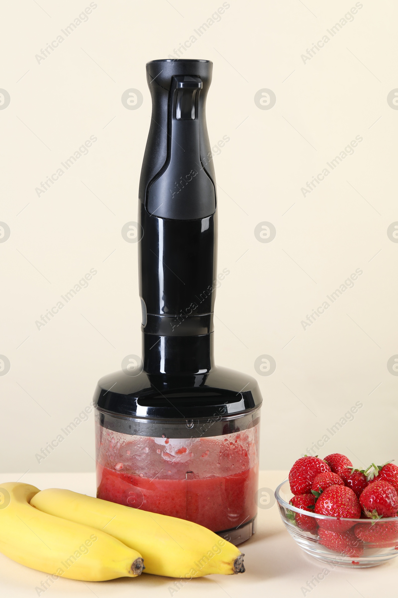 Photo of Hand blender with mixture of ingredients, banana and strawberries on beige background