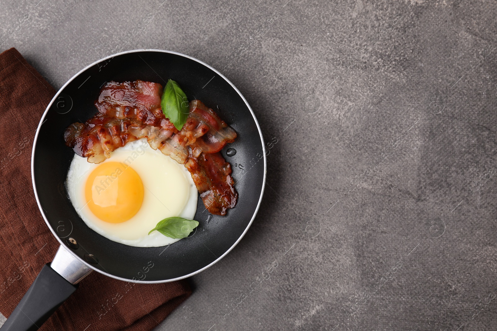 Photo of Fried egg, bacon and basil in frying pan on brown table, top view. Space for text