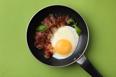 Photo of Fried egg, bacon and basil in frying pan on green background, top view