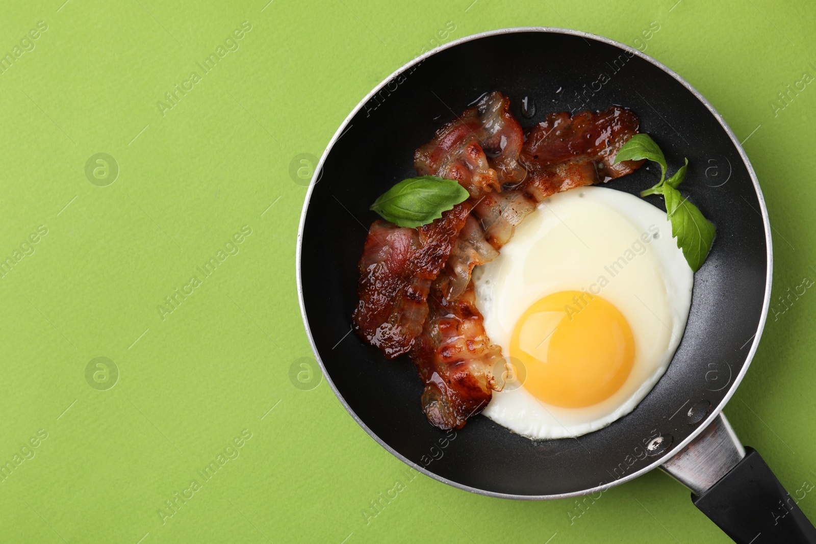 Photo of Fried egg, bacon and basil in frying pan on green background, top view. Space for text