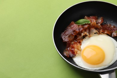 Photo of Fried egg, bacon and basil in frying pan on green background, space for text