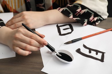 Photo of Calligraphy. Woman dipping brush into inkwell at wooden table, closeup. Papers with words Moon and Speak in Chinese