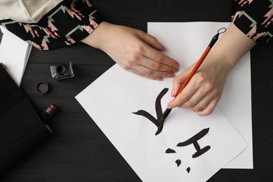 Calligraphy. Woman with brush and inkwell writing words River and Entrance in Chinese on paper at black wooden table, top view