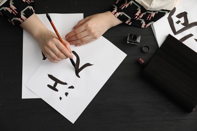 Calligraphy. Woman with brush and inkwell writing words River and Entrance in Chinese on paper at black wooden table, top view