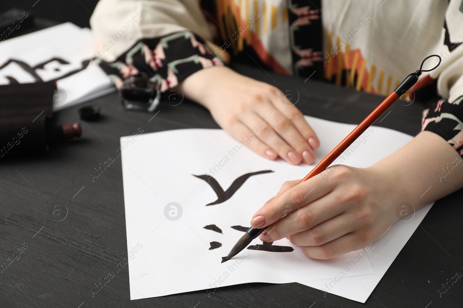 Photo of Calligraphy. Woman with brush writing words River and Entrance in Chinese on paper at black wooden table, closeup