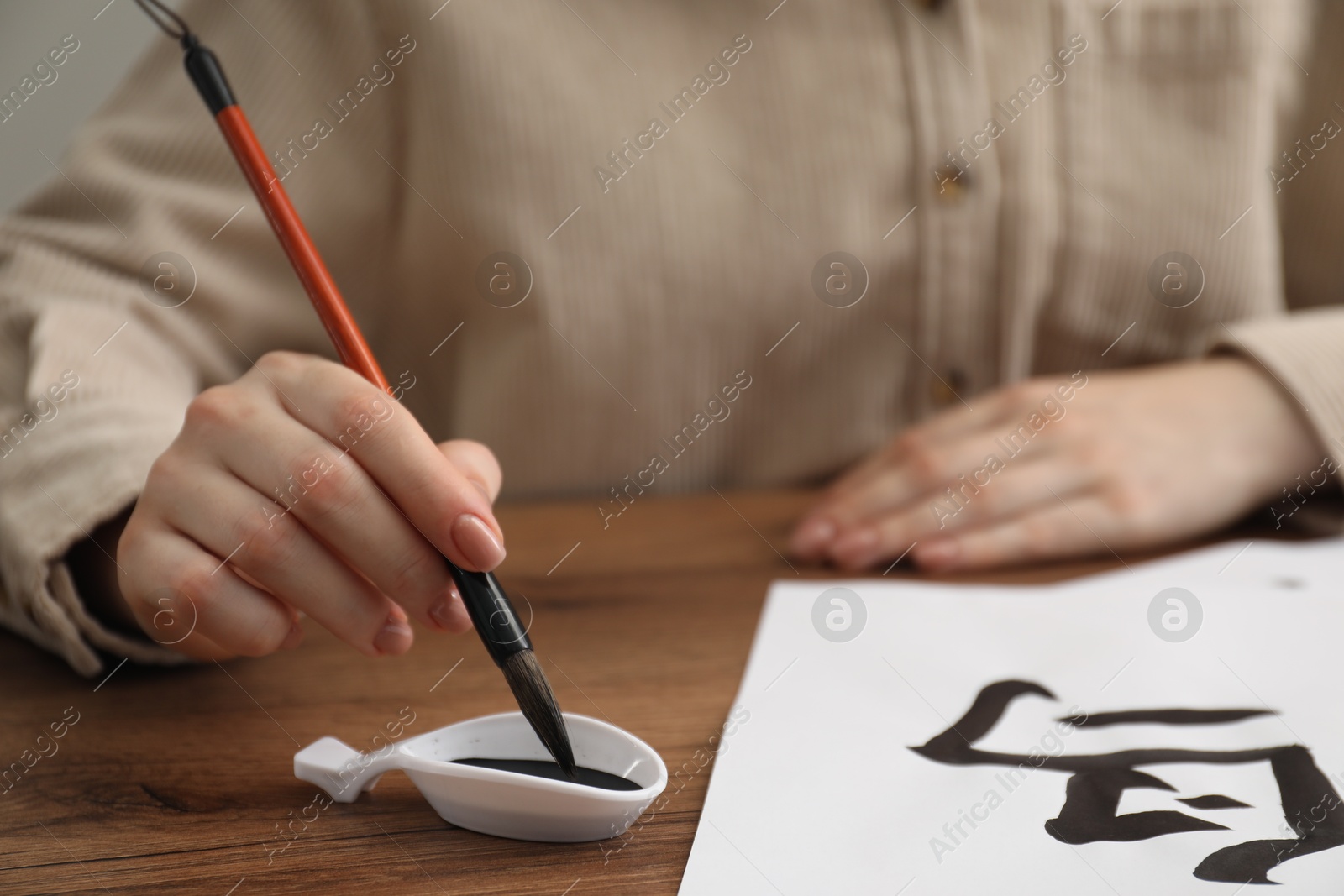 Photo of Calligraphy. Woman dipping brush into inkwell at wooden table, closeup. Practicing writing Chinese hieroglyphs