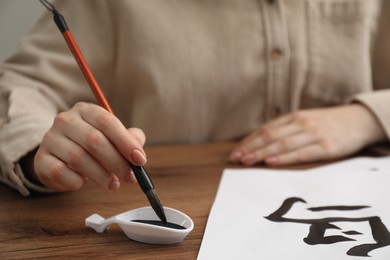 Calligraphy. Woman dipping brush into inkwell at wooden table, closeup. Practicing writing Chinese hieroglyphs