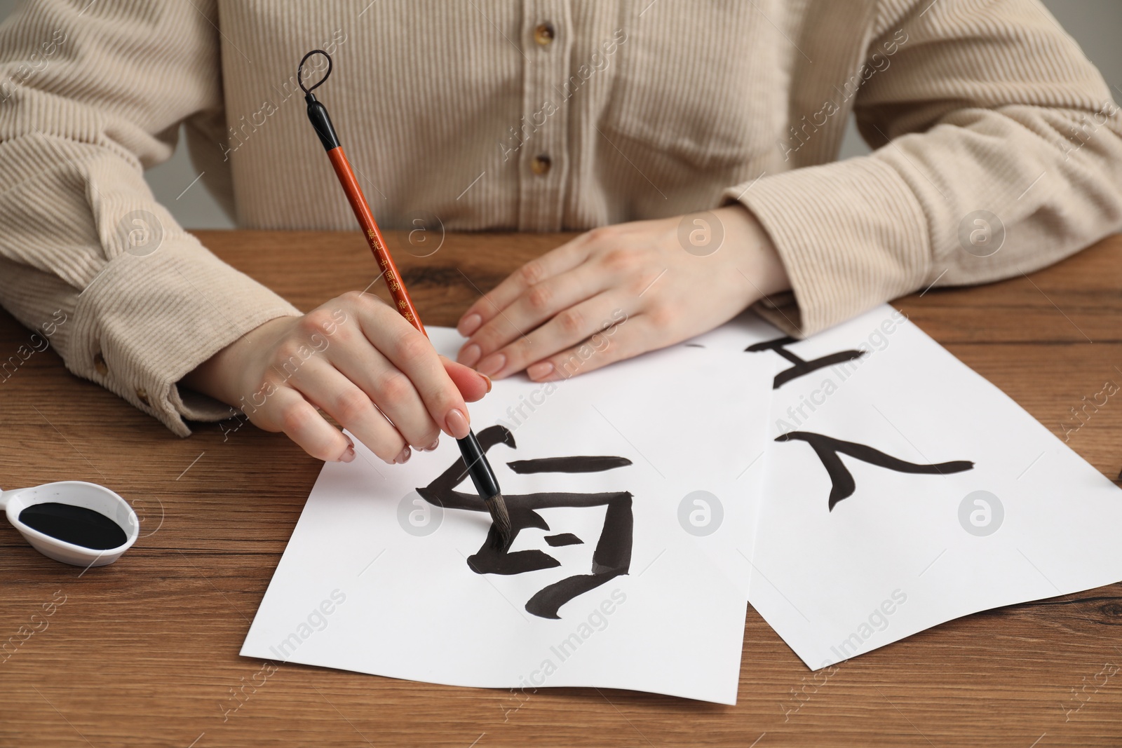 Photo of Calligraphy. Woman with brush and inkwell writing words Bird, River and Entrance in Chinese on paper at wooden table, closeup