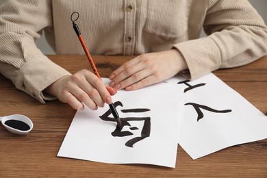 Calligraphy. Woman with brush and inkwell writing words Bird, River and Entrance in Chinese on paper at wooden table, closeup